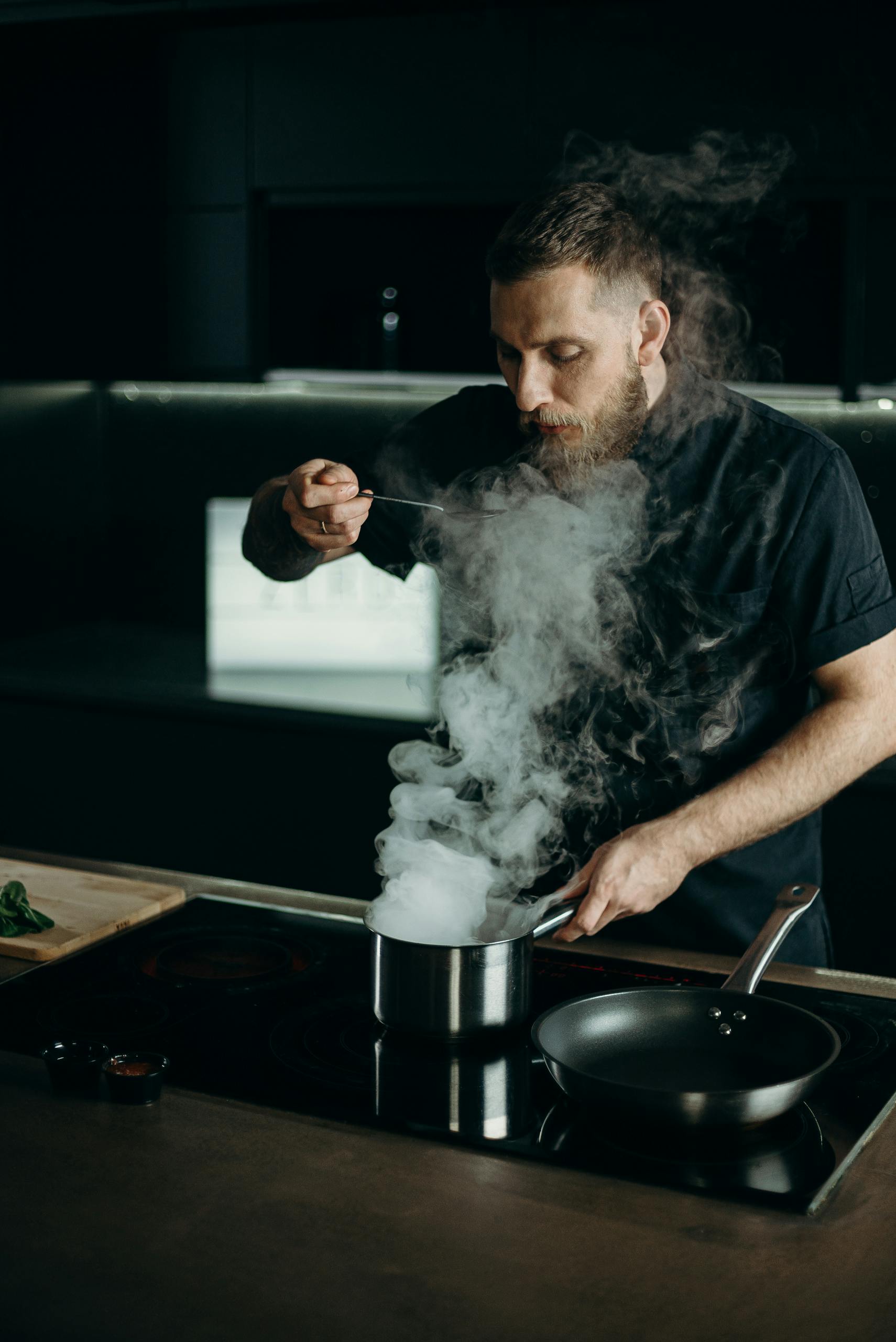 A professional chef taste-testing a dish with steam rising from the pot in a modern kitchen.